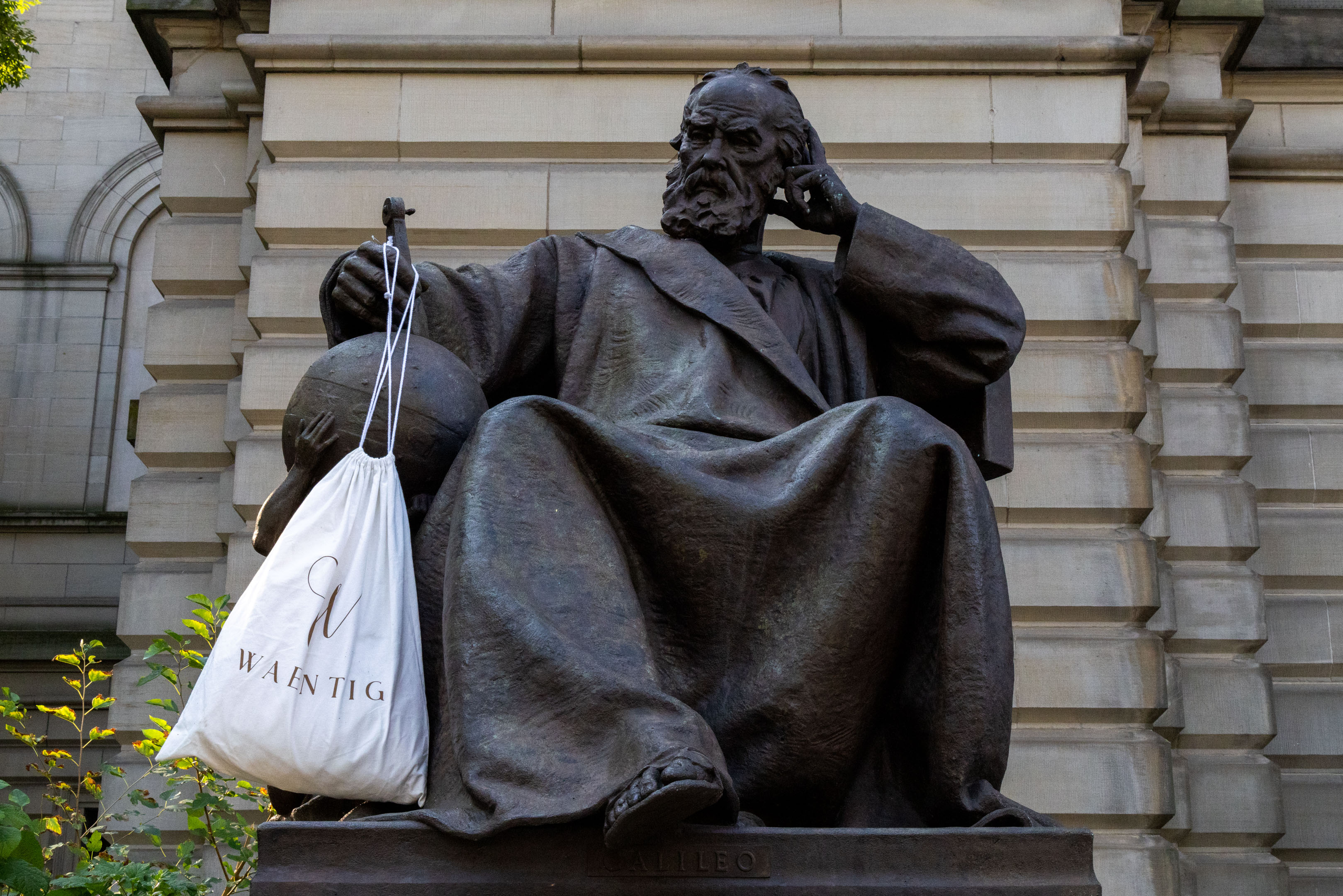 a statue of Galileo sitting with his legs crossed with a white canvas bag in his hand